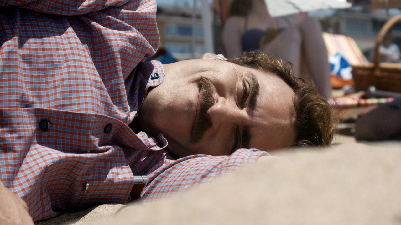 Ein Mann mit Schnurrbart liegt am Strand, lächelt sanft und ruht seinen Kopf auf dem Arm. Im Hintergrund sind weitere Personen und Strandstühle zu sehen. Das Bild zeigt den Hauptprotagonisten Theodore aus dem Film HER, der seine KI-Companion Sam anschaut.
