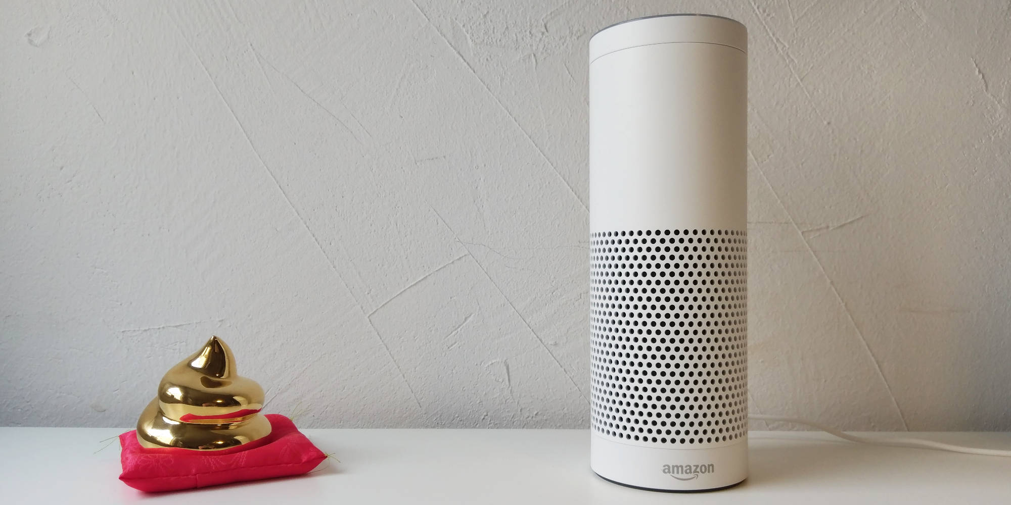 Eine weiße Alexa, der Smart Speaker mit Voice User Interface von Amazon, steht auf einer weißen Fläche neben einer kleinen goldenen Skulptur in Form eines Kackhaufens. Die Skulptur ruht auf einem roten Kissen vor einer schlichten hellgrauen Wand.